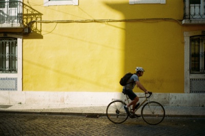 Street photograph of a cyclist passing a yellow wall in Lisbon, harsh afternoon shadows, 35mm film, generated at 3:2