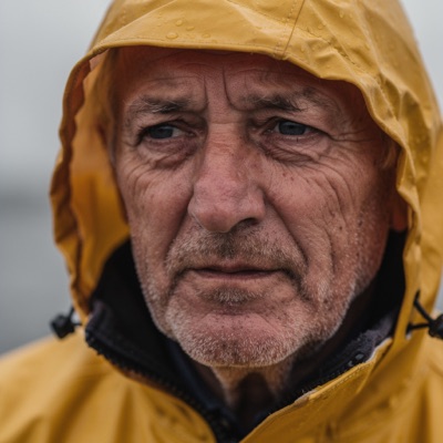 Close-up portrait of an elderly fisherman with a weathered face and grey stubble, wearing a yellow raincoat, soft overcast light from a window, shallow depth of field, 85mm photograph, generated by FLUX.2 [pro]