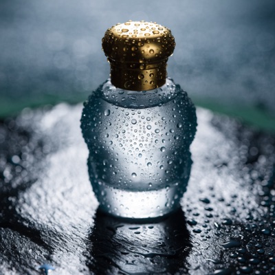 A frosted glass perfume bottle with a brass cap on wet black slate, droplets on the glass, soft rim light, reflections on the stone, studio product photography, generated by Recraft V4