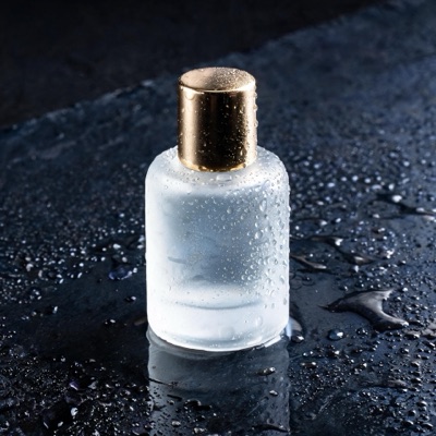 A frosted glass perfume bottle with a brass cap on wet black slate, droplets on the glass, soft rim light, reflections on the stone, studio product photography, generated by Seedream 5 Lite